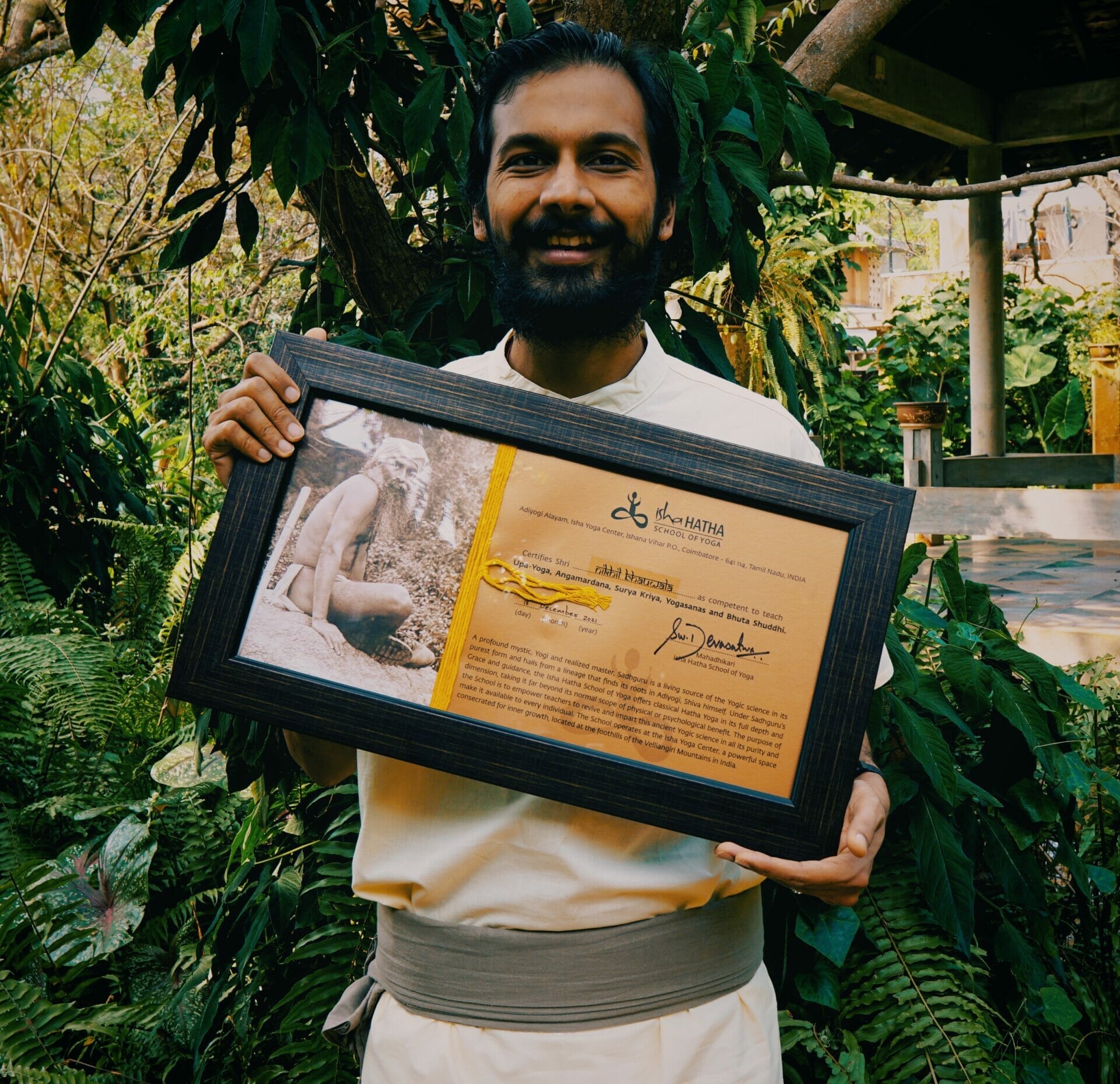 A yoga practitioner, Isha, celebrates his achievement in India by holding a framed certificate in front of a tree.