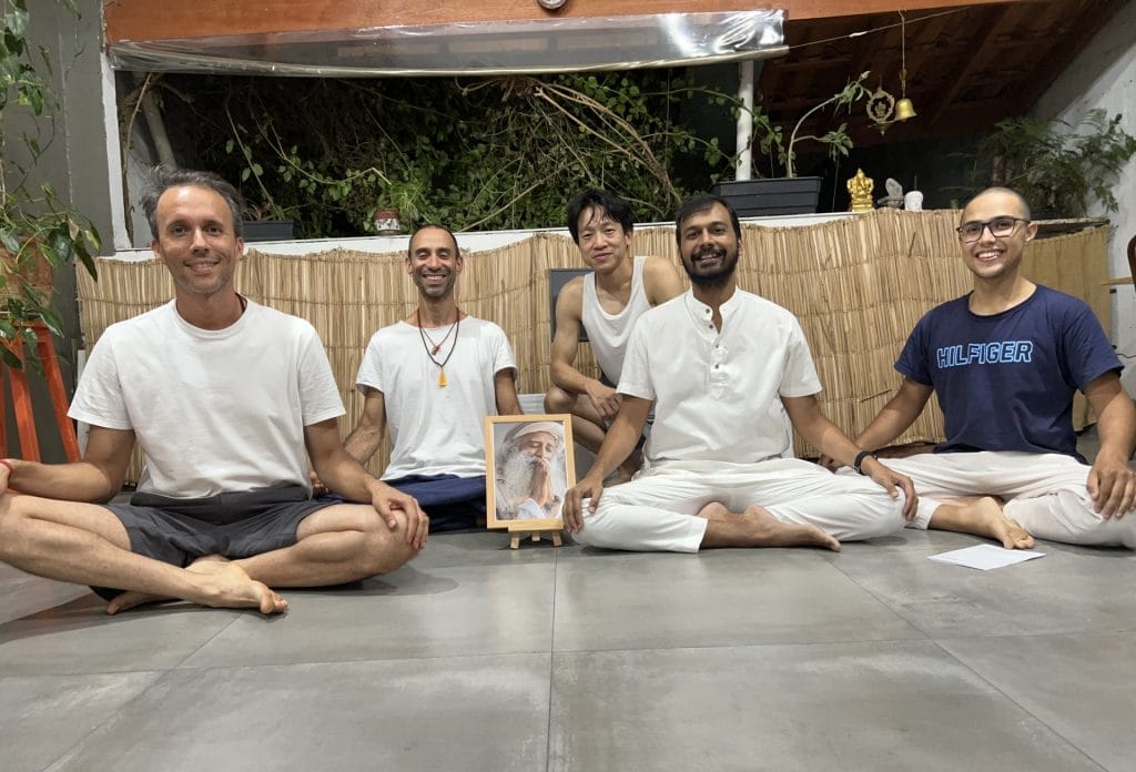 Five people in casual and traditional clothing sitting cross-legged on mats in a yoga studio, smiling at the camera.