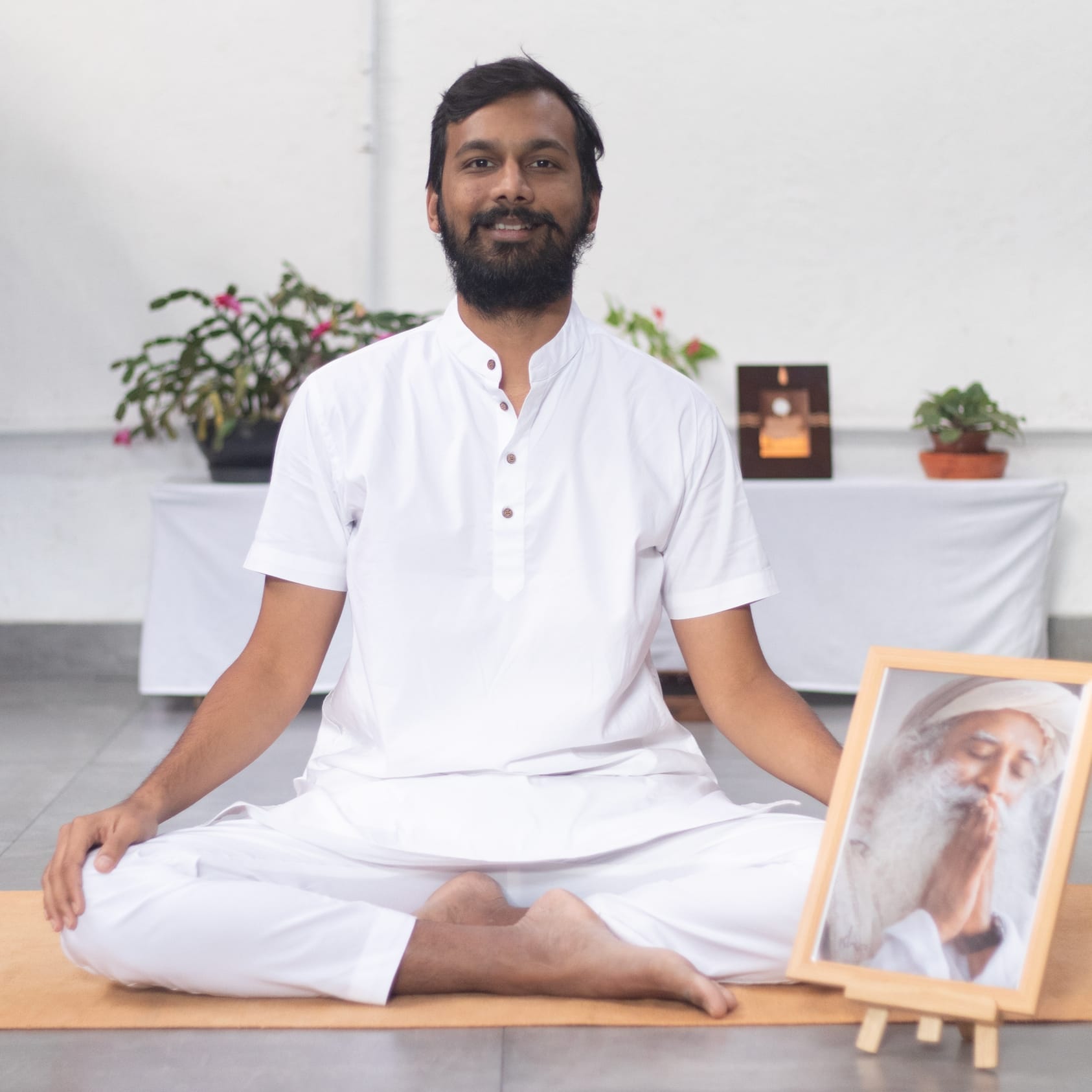 A person in white clothing sits cross-legged on a yoga mat, a framed photograph of Sadhguru beside them. Plants and items are artfully arranged on a table in the background.