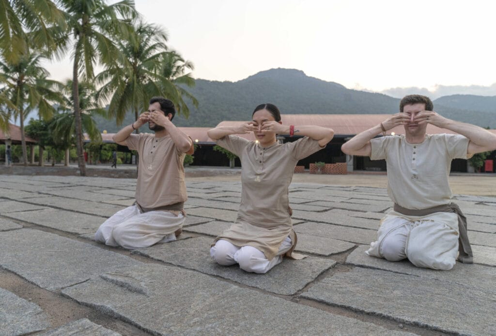 Three individuals in light-colored clothing kneel outdoors on stone tiles, covering their ears while facing forward. Palm trees and a mountainous backdrop are visible in the background, reminiscent of serene yoga retreats in India.