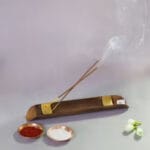 Two incense sticks burning on a bronze holder with smoke rising. Nearby are small bowls with red and white powders and a few white flowers.