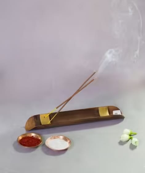 Two incense sticks burning on a bronze holder with smoke rising. Nearby are small bowls with red and white powders and a few white flowers.