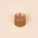 A small, round, open-top metal container with a copper finish on a light beige background.