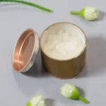 Open gold tin with white cream inside on a gray surface, surrounded by small white flowers and green stems.