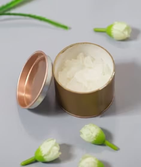 Open gold tin with white cream inside on a gray surface, surrounded by small white flowers and green stems.