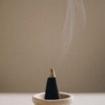 A smoking incense cone in a small ceramic holder on a light surface, with smoke gently rising against a neutral background.